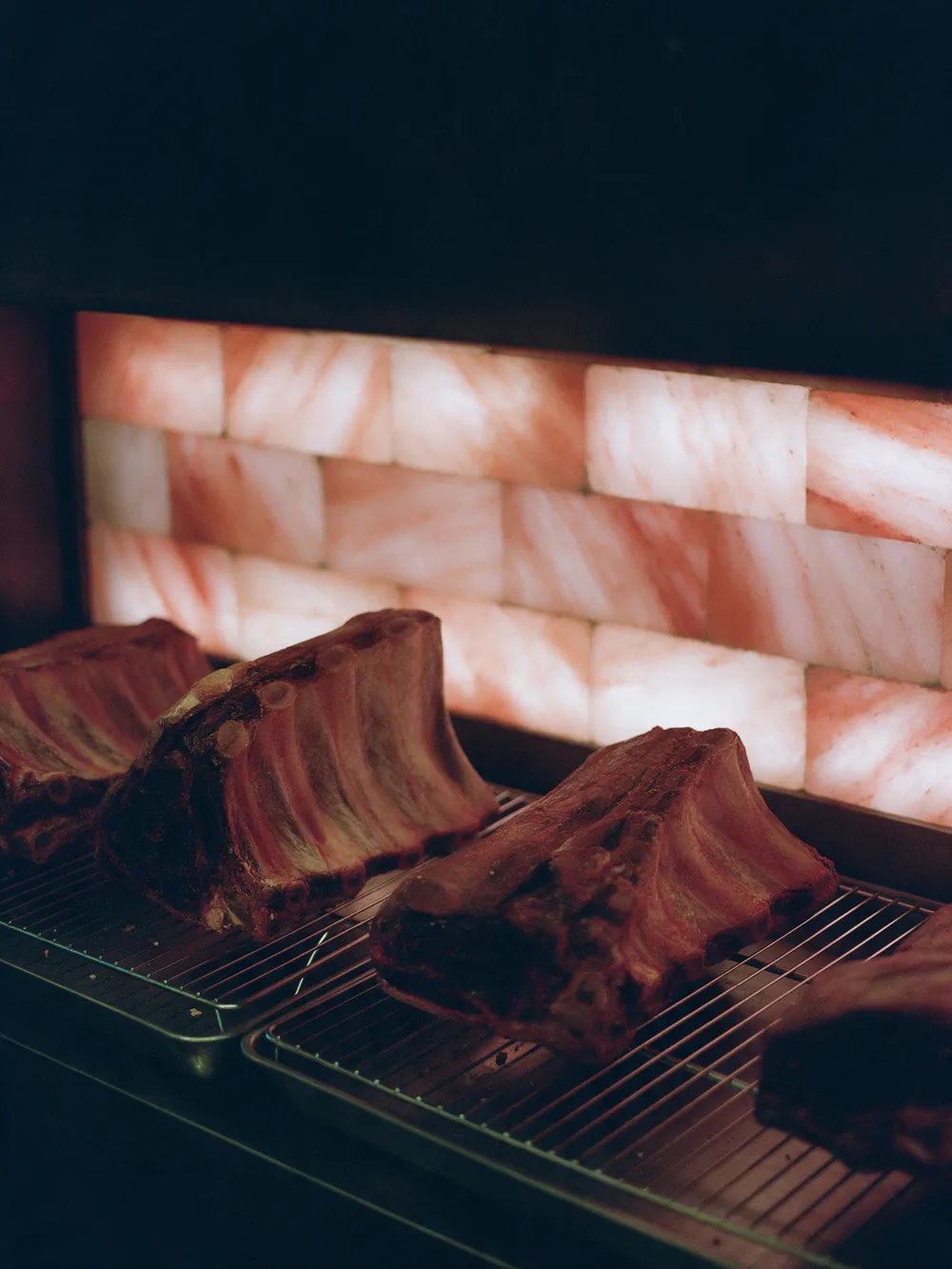 Inside the dry-ageing room: whole beef ribs, dark with age, on steel racks in front of a glowing wall of pink salt bricks.