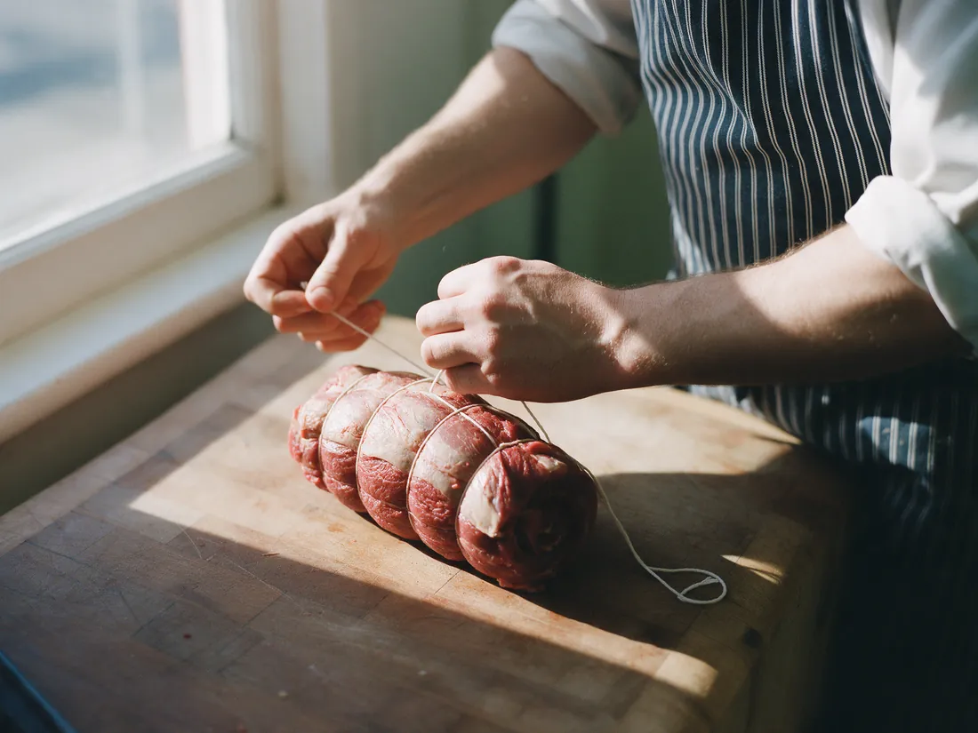 A butcher's hands tying a rolled beef roast with white twine on a worn timber bench, afternoon light from the window.