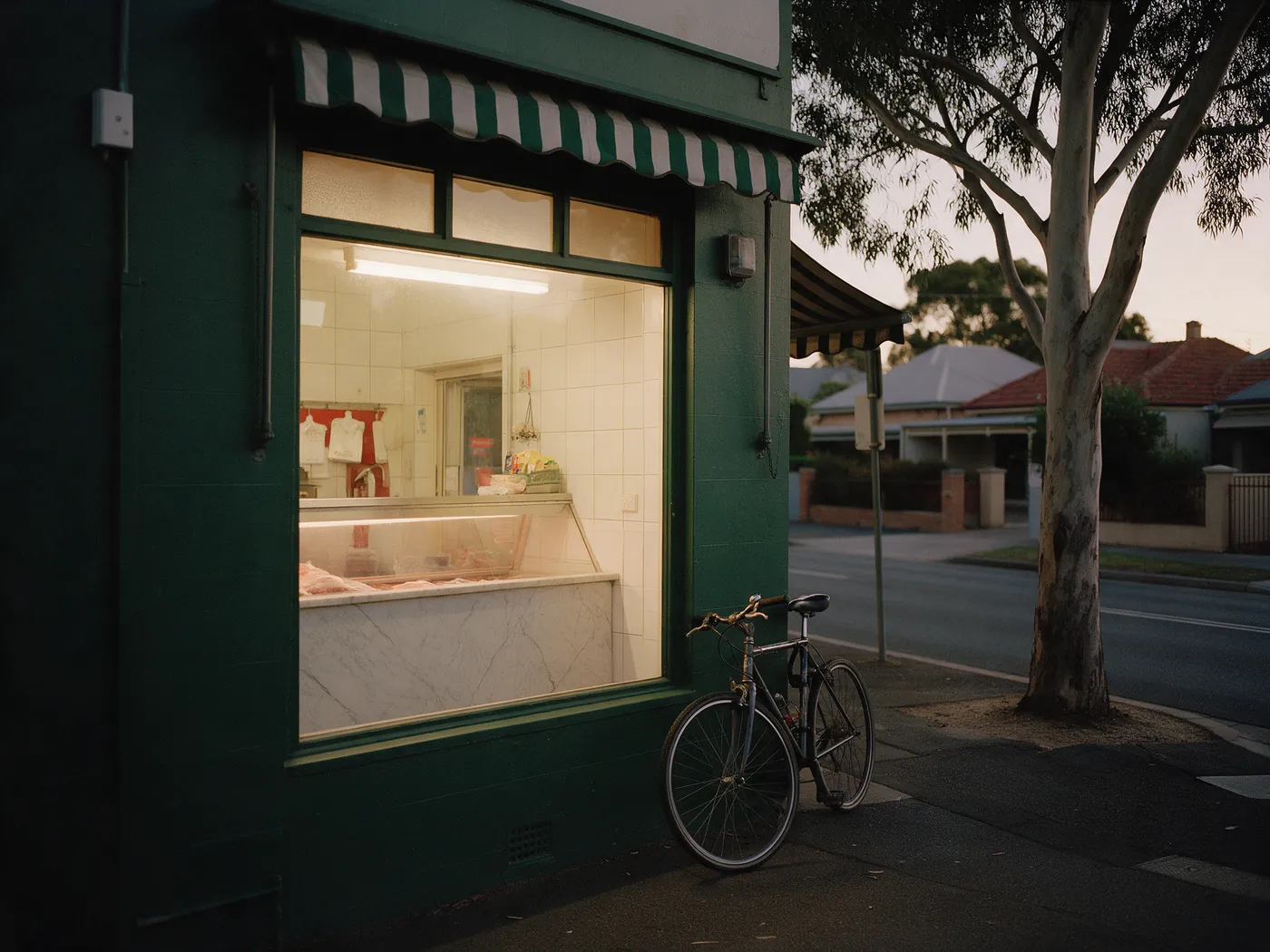 A bottle-green butcher's shopfront at dusk, its window glowing warm, a bike against the wall and a gum tree at the kerb.